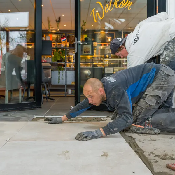 Twee tegelzetters leggen grote vloertegels bij de ingang van een winkel